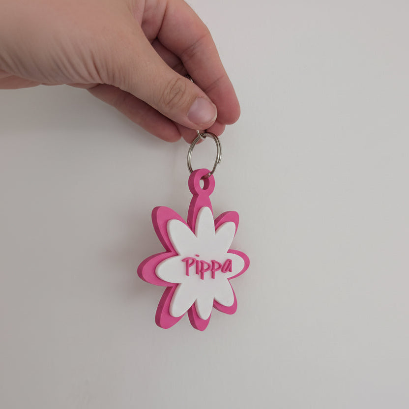 Hand holding a pink and white flower-shaped keychain with 'Pippa' engraved on a light gray background