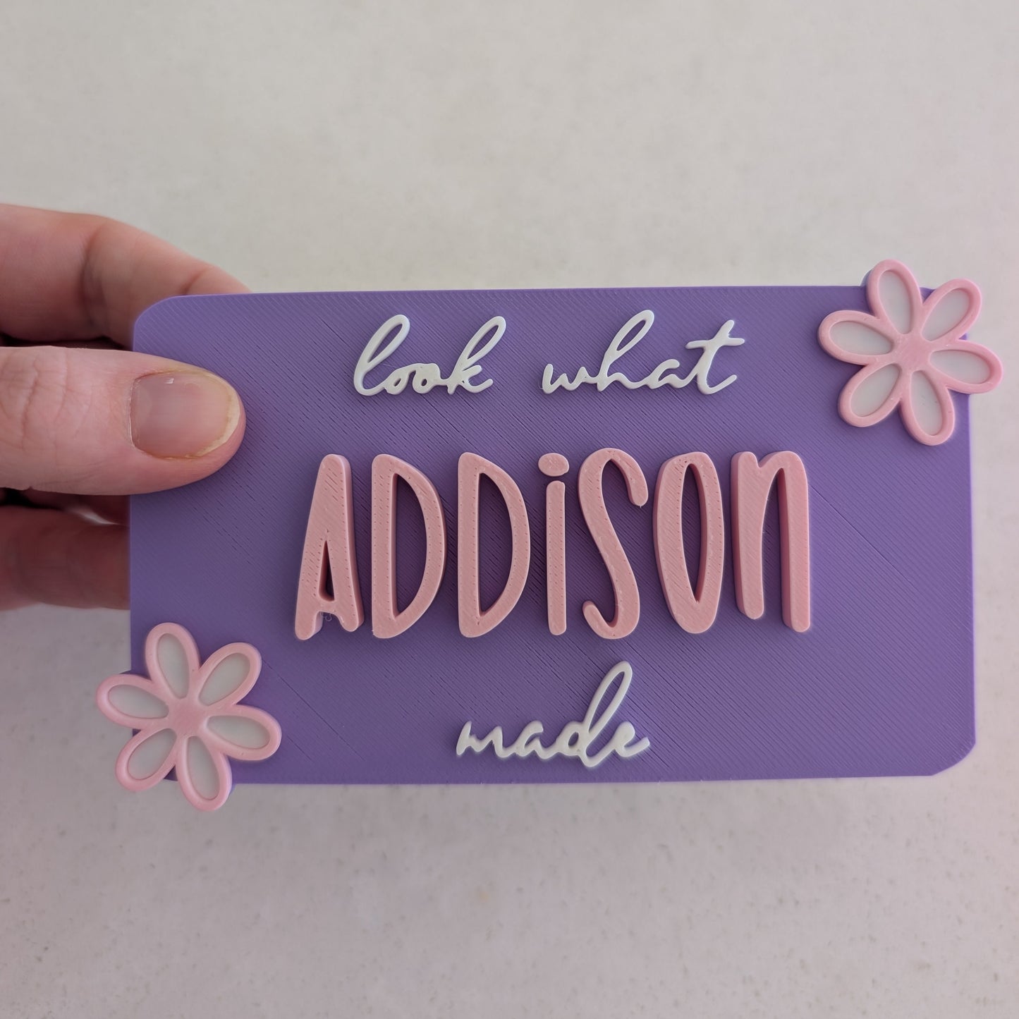 Purple magnet with 'look what Addison made' text and pink flowers held by a hand on a light gray background