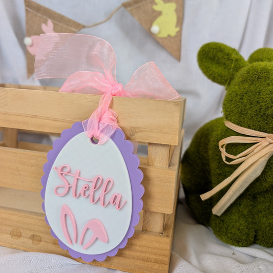 Wooden crate with a decorative tag and pink bow, next to a green plush toy, on a light background.