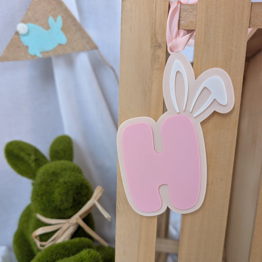 Pink letter 'H' with bunny ears on a wooden stand, surrounded by Easter decorations.
