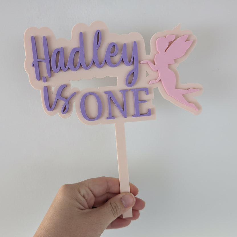 Hand holding a cake topper with 'Hadley is one' and a fairy design on a white background