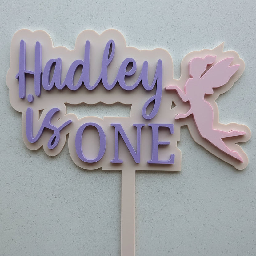 Cake topper with 'Hadley is ONE' text and a fairy design on a light gray background