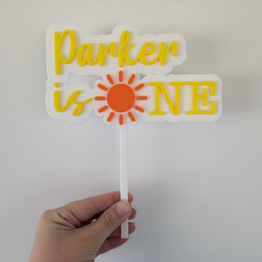 Hand holding a decorative cake topper with 'Parker is ONE' text and sun design.