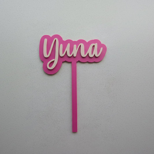 Pink cake topper with 'Yuna' on a light gray background